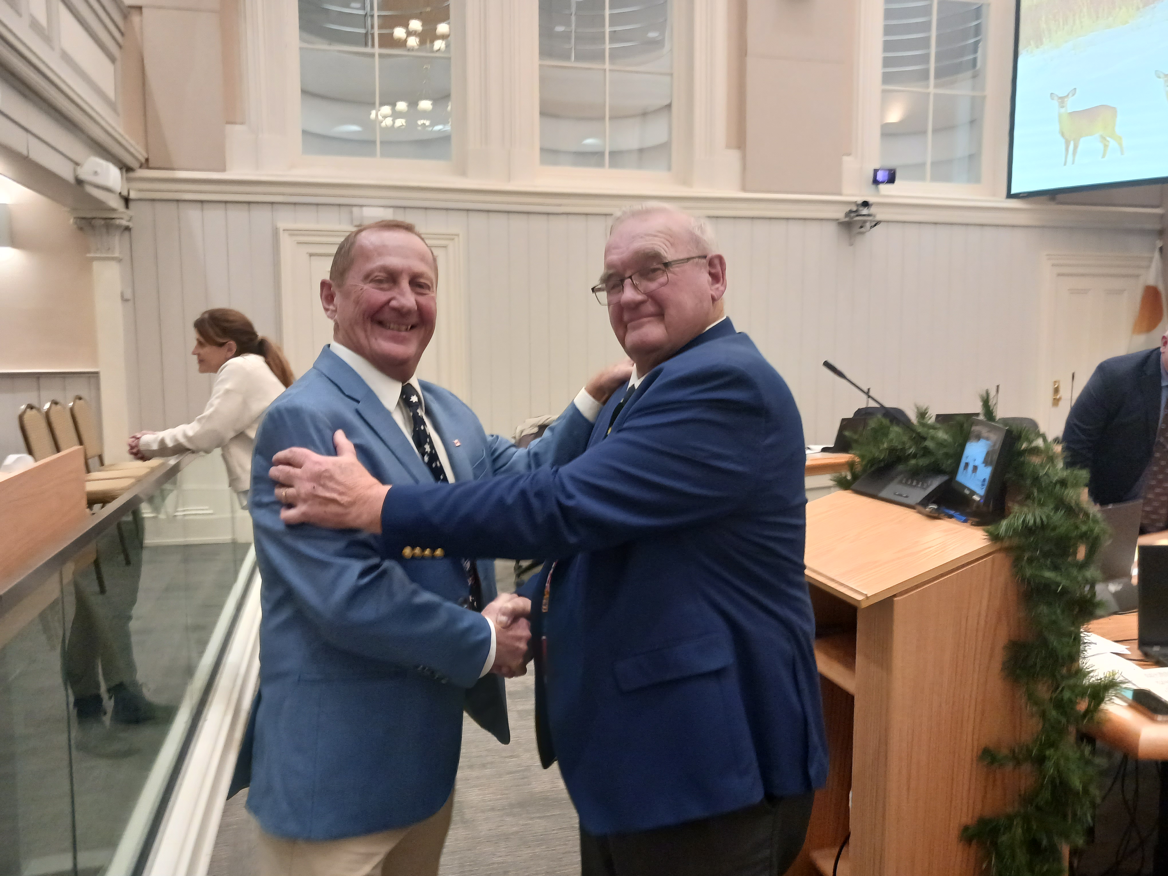 Two men wearing blue blazers shake hands and smile.