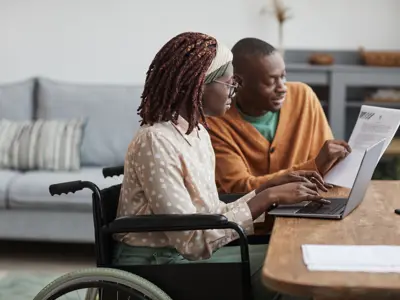 Two people sitting at table looking at laptop and piece of paper.
