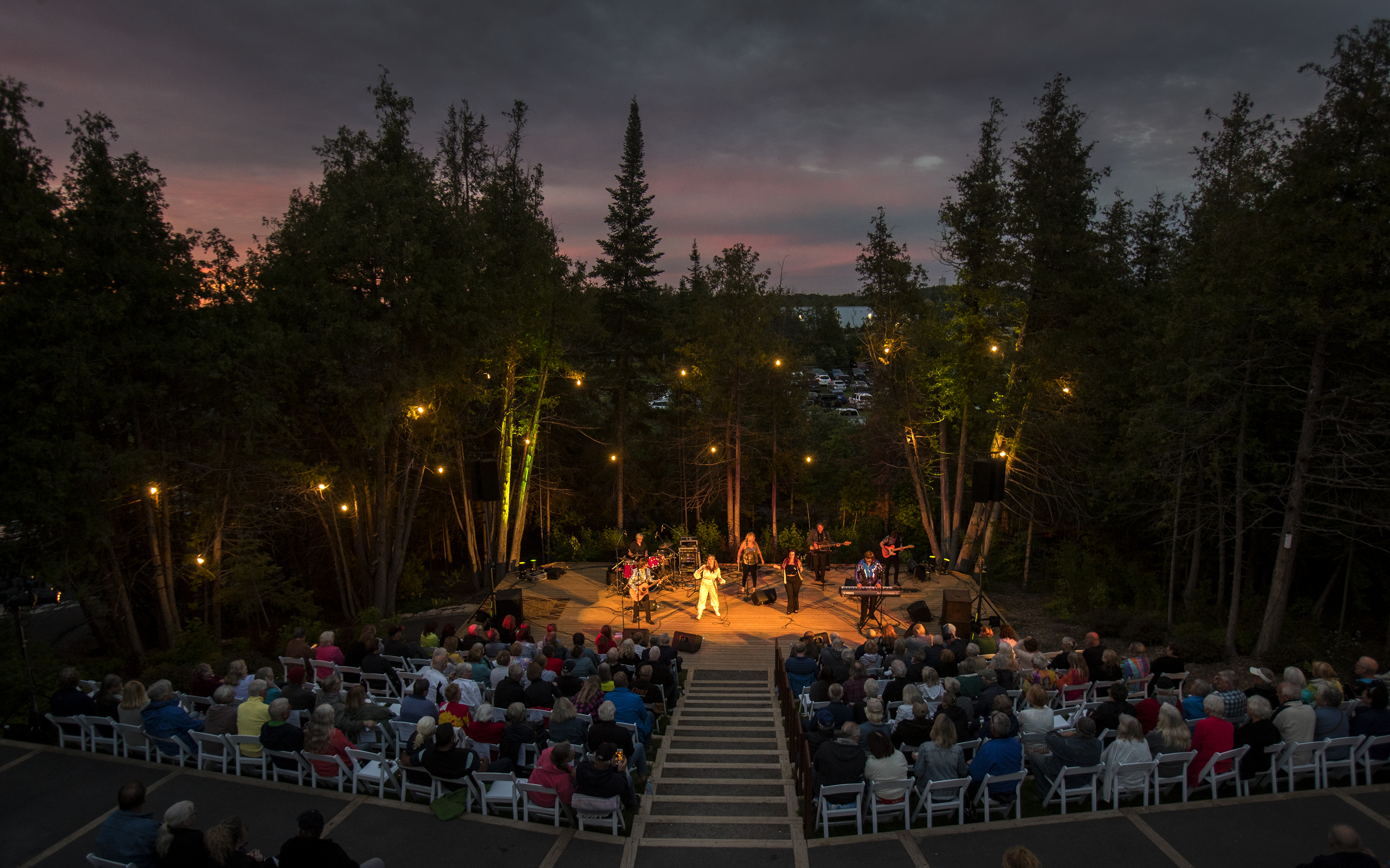 A lit outdoor stage, surrounded by trees is watched by a full audience.