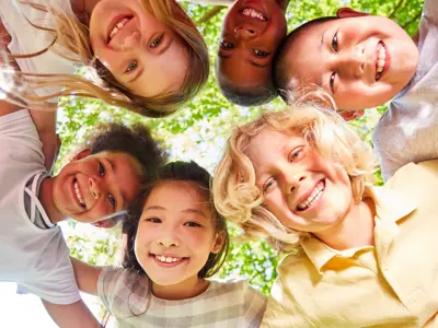 Photo of six children leaning over a camera and smiling. Their arms are around each other, and they are a mix of genders, skin tones and ethnicities.
