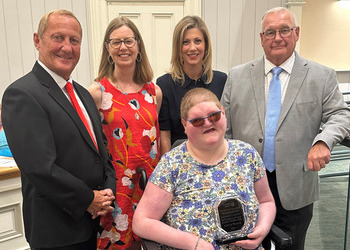 A young woman using a wheelchair sits holding an award. Two men and two women stand behind her.