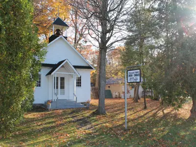 Photo of a white clapboard church with a steel bell in the small steeple. A sign in front reads 'Pleasant Point Union Church.' The ground around the church is grassy and dotted with orange and brown leaves that have fallen.