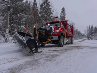 A red snowplow is moving snow off the side of a road on a cloudy winter day