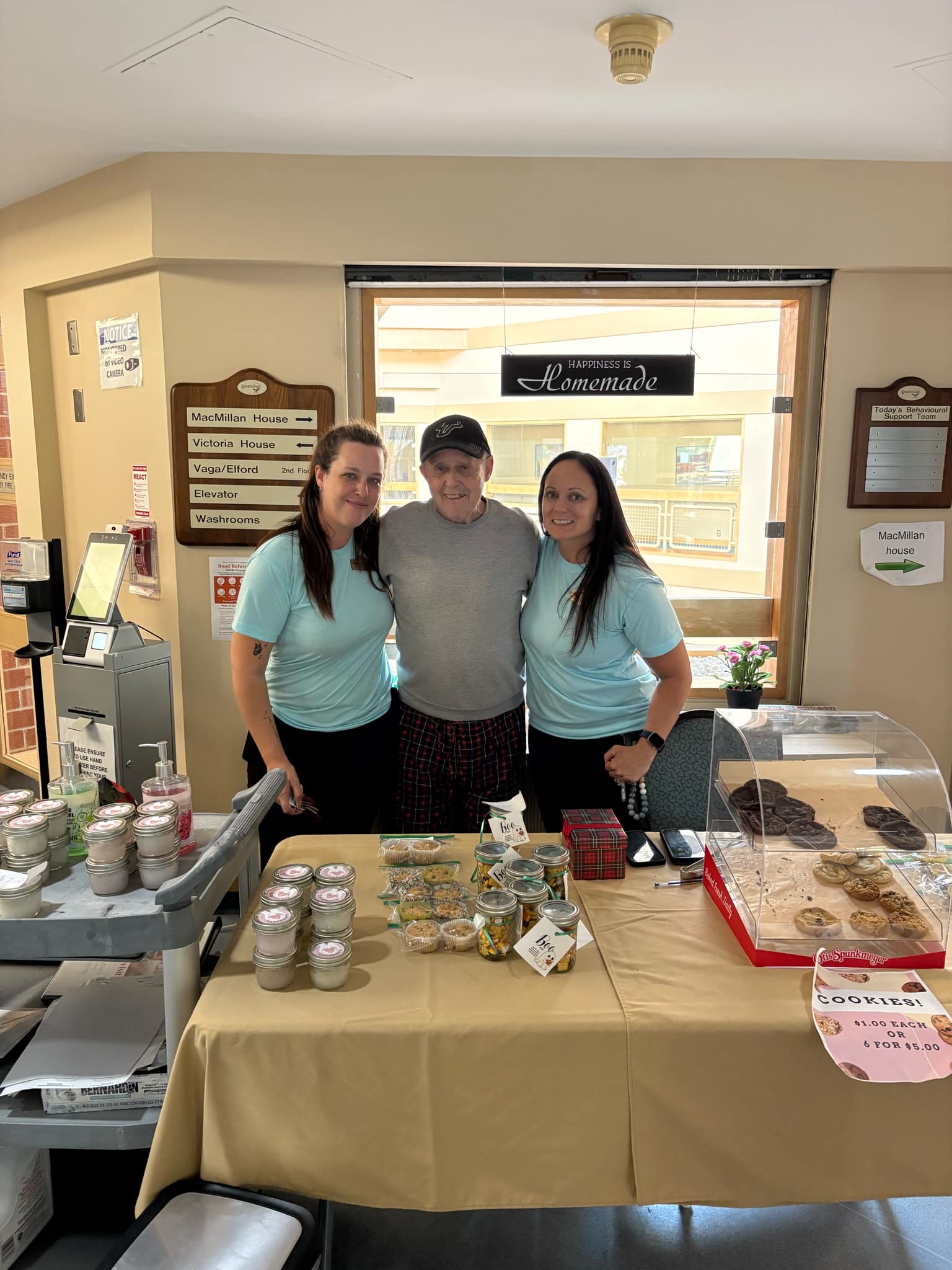 Two women and an older man stand behind a table of baked goods.