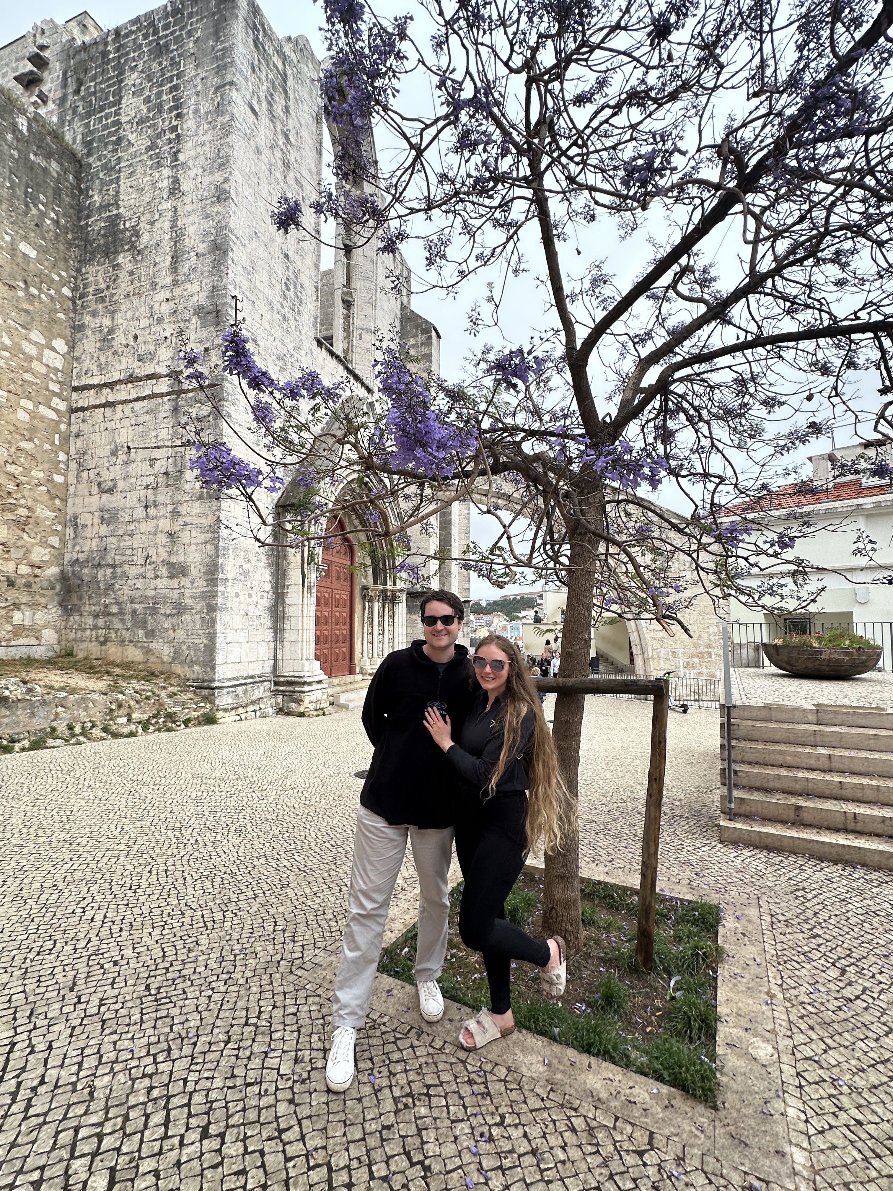 A man and women stand under a tree blooming with purple flowers. An old stone church is in the background, and the ground is paved with cobblestones.