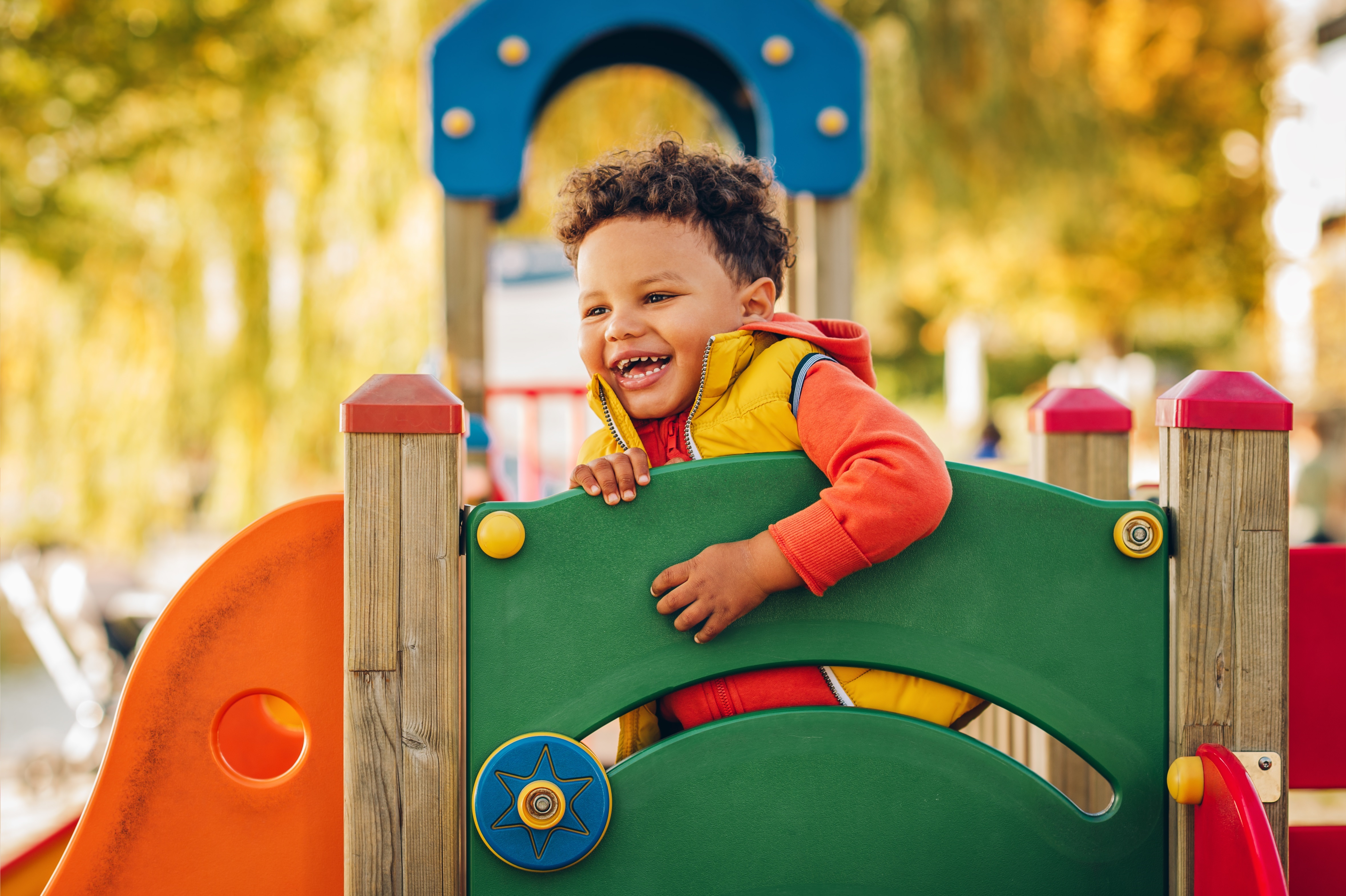 A child with curly brown hair plays on colourful playground equipment. Leafy trees are in the background.