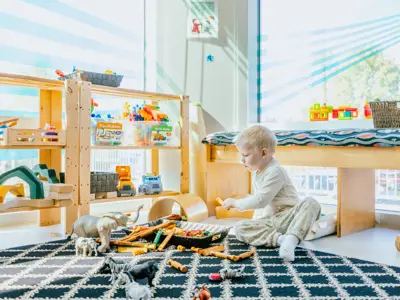 Photo of young child playing on the floor, surrounded by shelves of toys and windows.