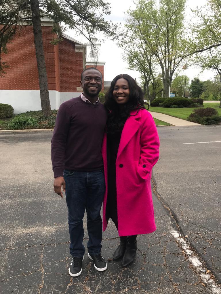 A man in a dark sweater stands with his arm around a woman in a long fuscia coat. They are both smiling and there is a red brick building in the background.