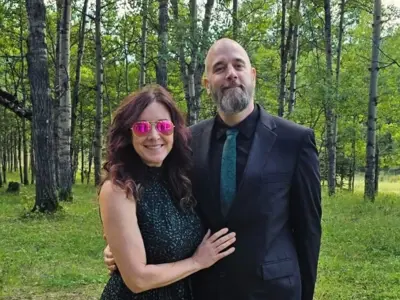 A woman in a black dress and sunglasses with a man in a black suit and tie pose and smile for the camera. The background shows a forest of leafy green trees and grasses.