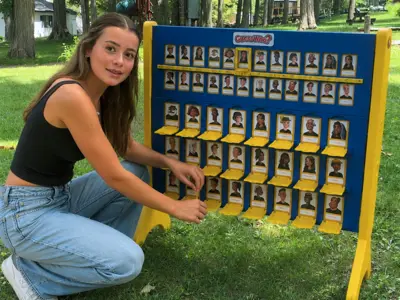A young woman is crouched down beside a lawn game on the grass, with trees in the background.