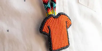 Photo of an Indigenous beaded necklace, with a beaded orange shirt pendant. A white button-up shirt is the background.