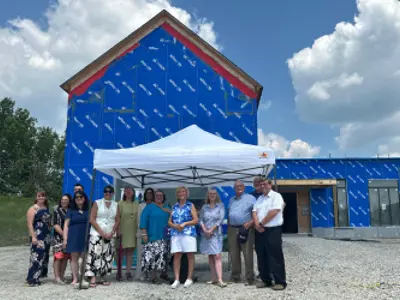 A group of officials stand in front of a building under construction. The building is wrapped in a blue construction material.