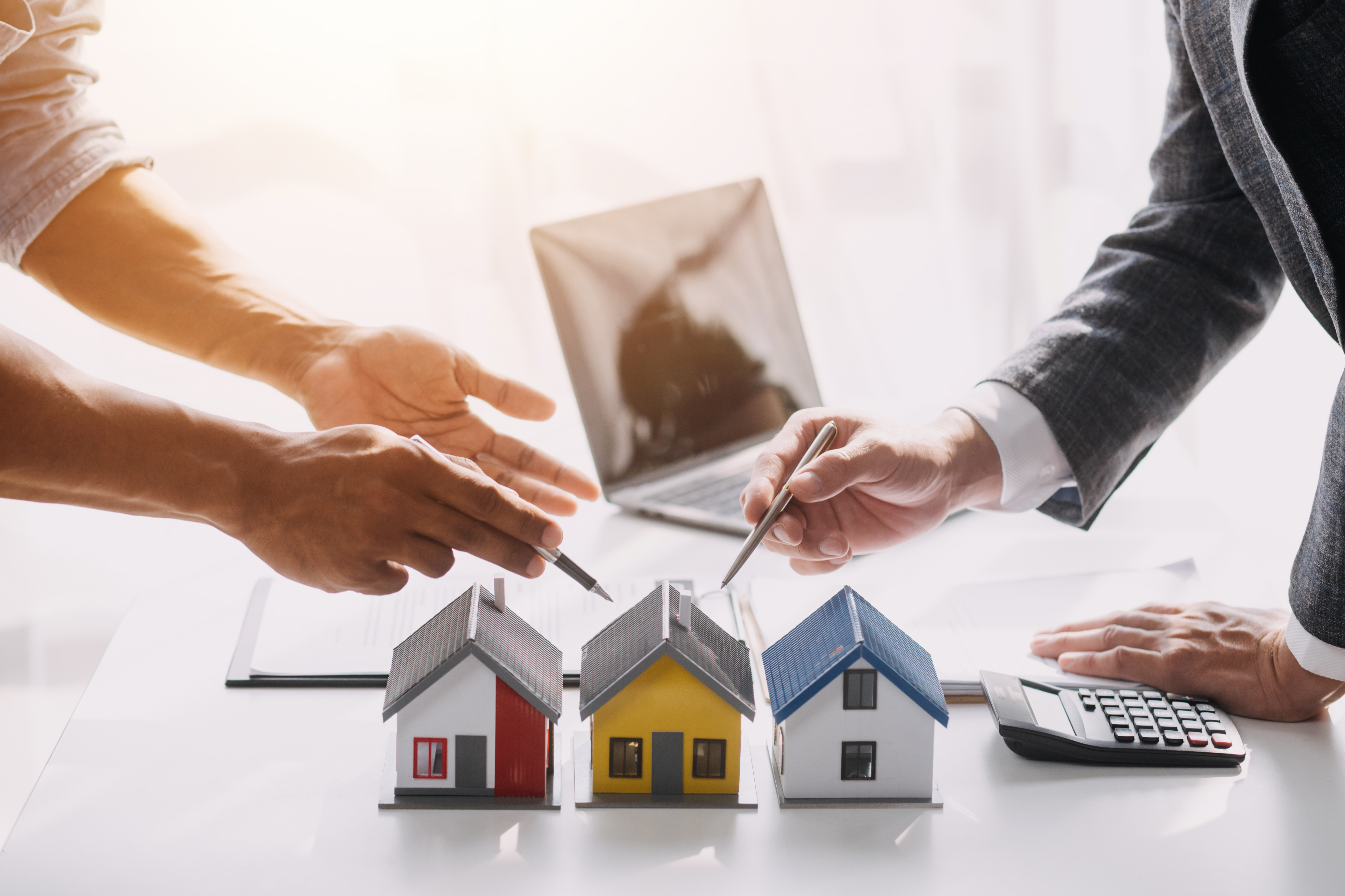 Three model houses on table with two people's hands gesturing with pens above the houses.