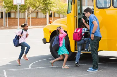 3 children in a line getting on a school bus while a teacher high fives the middle child.