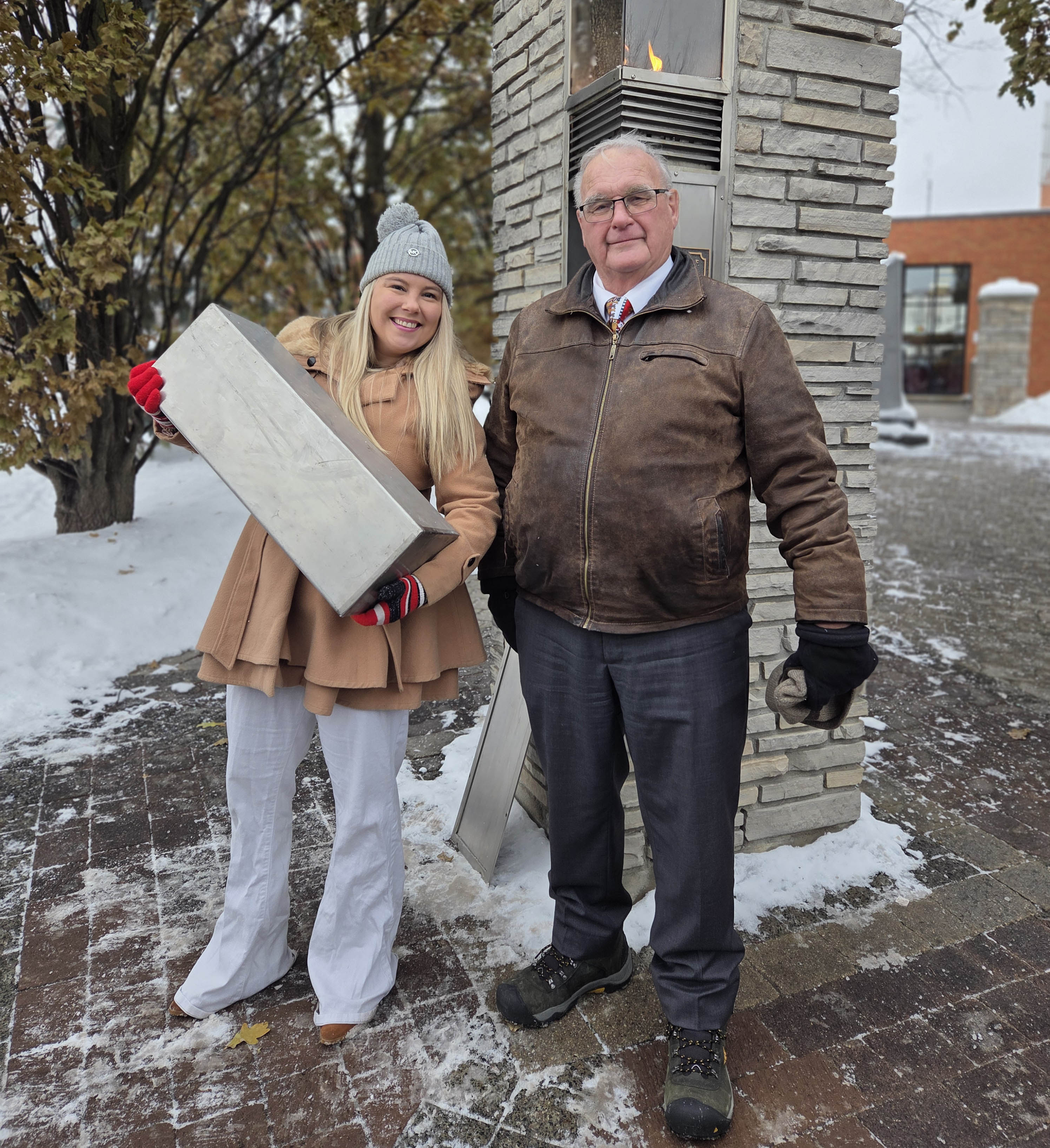 Photo of a young woman and a man standing in front of a brick pillar with a gas fire burning in it. The woman is holding a rectangular steel box.